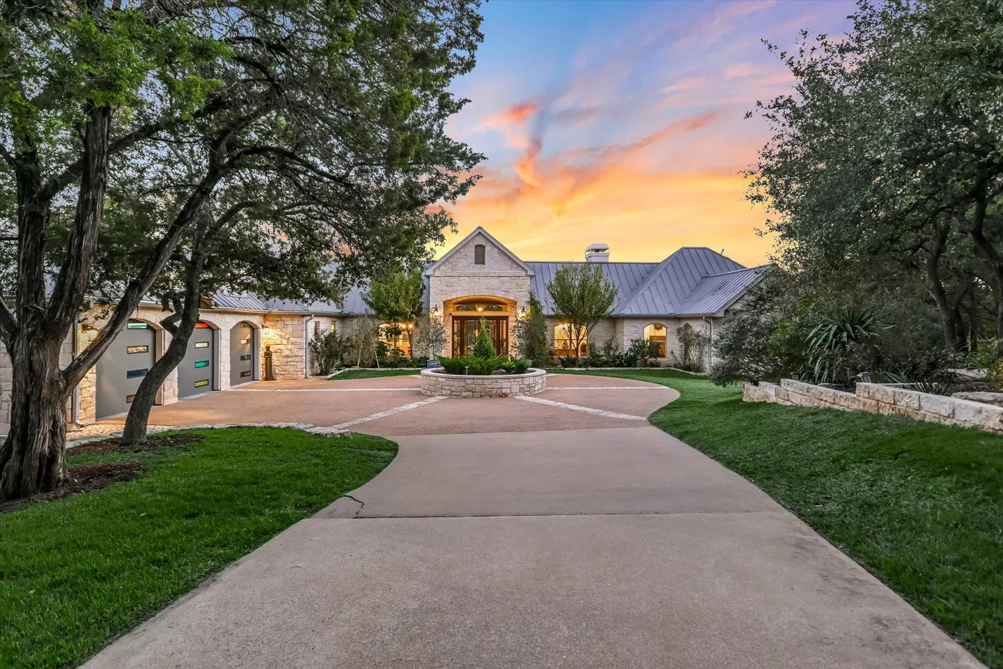 Exterior shot of a stone house with a long driveway, green lawn, and trees under a colorful sunset sky.