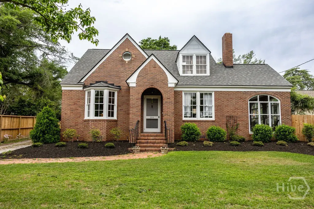 Brick house with gray roof, white trim, and green lawn. Bushes line the front of the house.