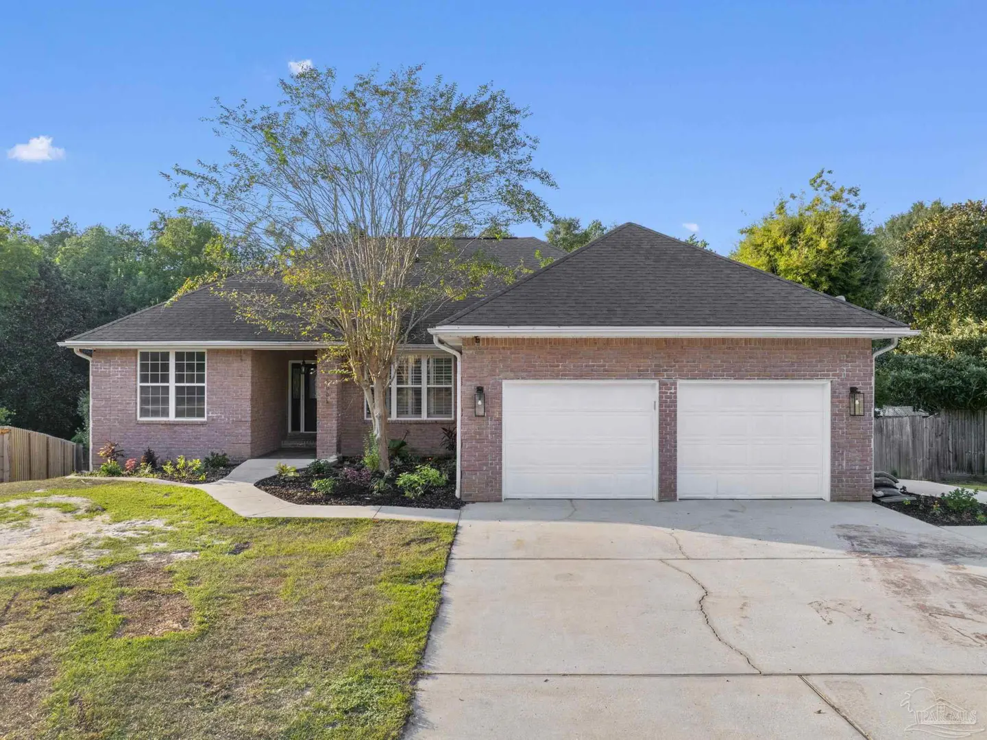 A single-story brick house with a gray roof, white garage doors, and a tree in the front yard under a blue sky.