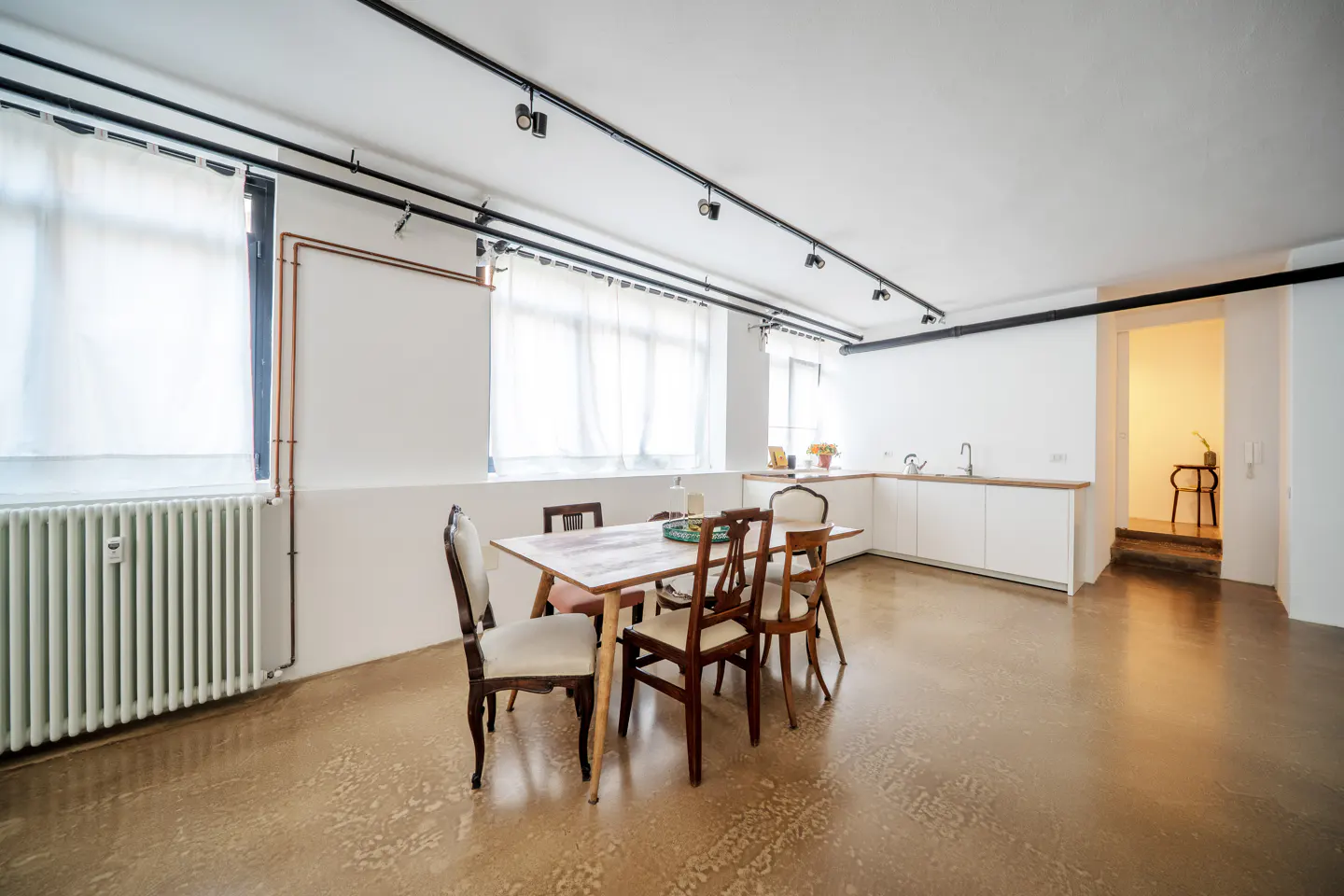 Bright, open-concept apartment with a dining table, chairs, and a white kitchen. Track lighting on the ceiling. Polished concrete floors.