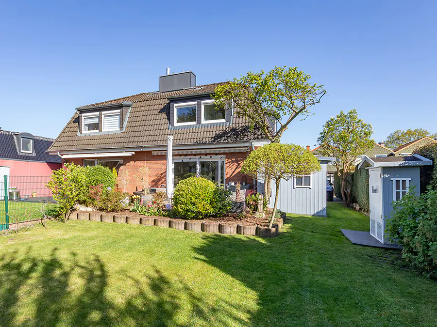 Backyard view of a brick house with a brown roof, green lawn, and blue sheds on a sunny day.