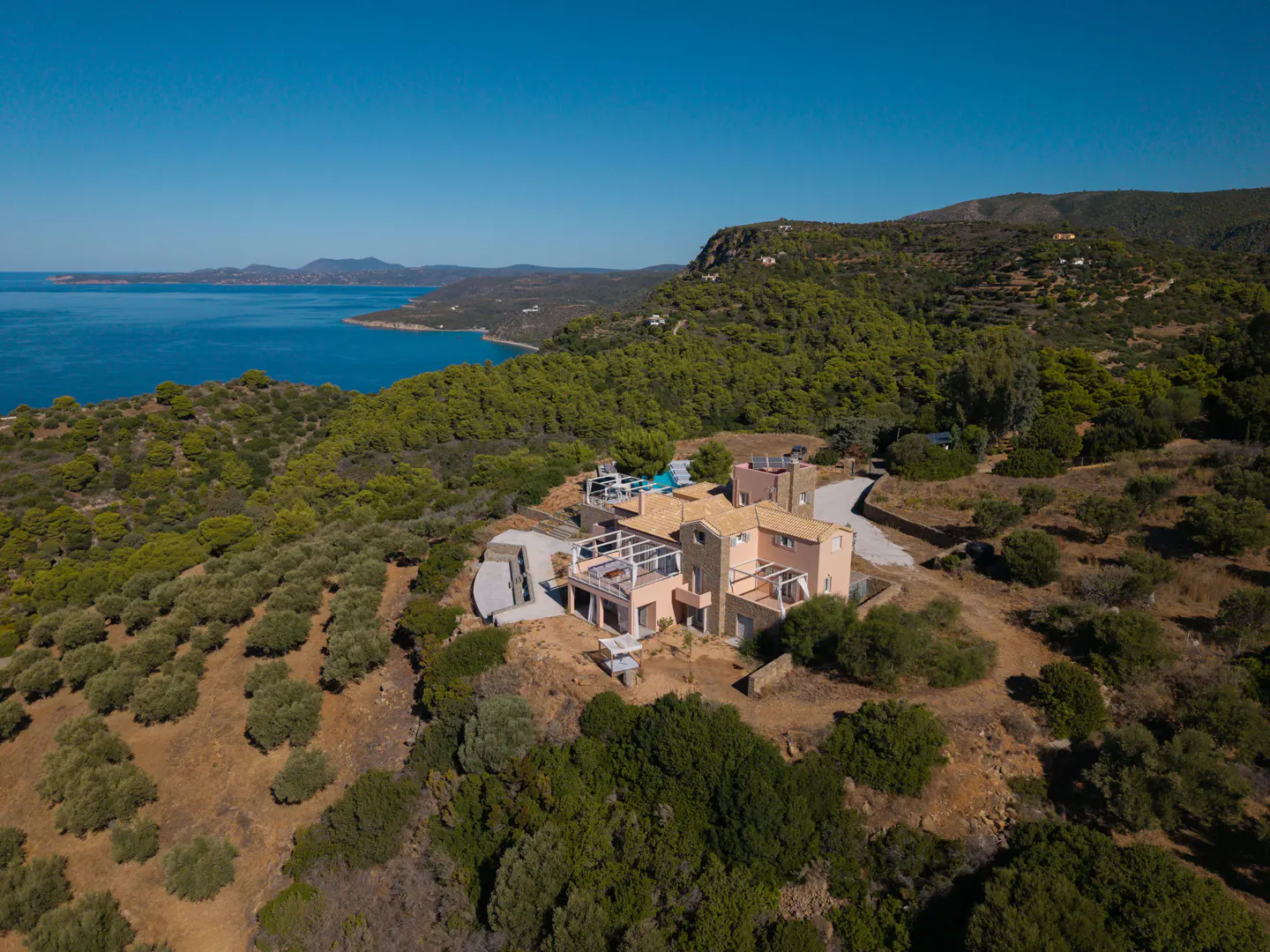 Aerial view of a peach-colored house with a blue pool, surrounded by green trees, with the ocean and blue sky in the background.