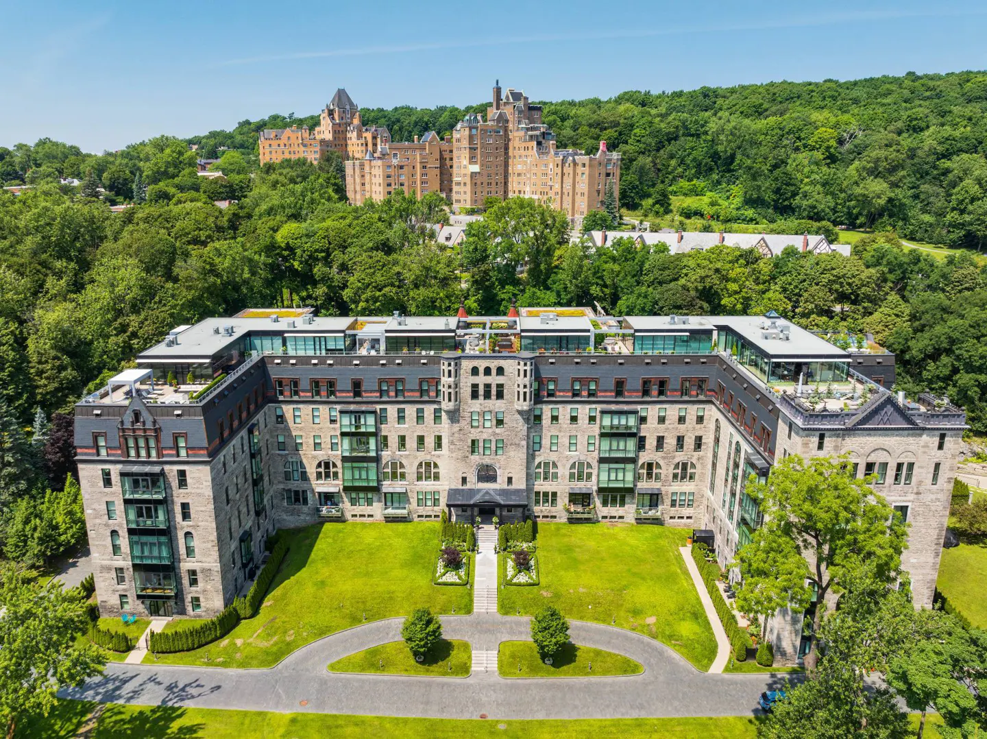 Aerial view of a stone apartment building with green lawn and trees, and a castle in the background.