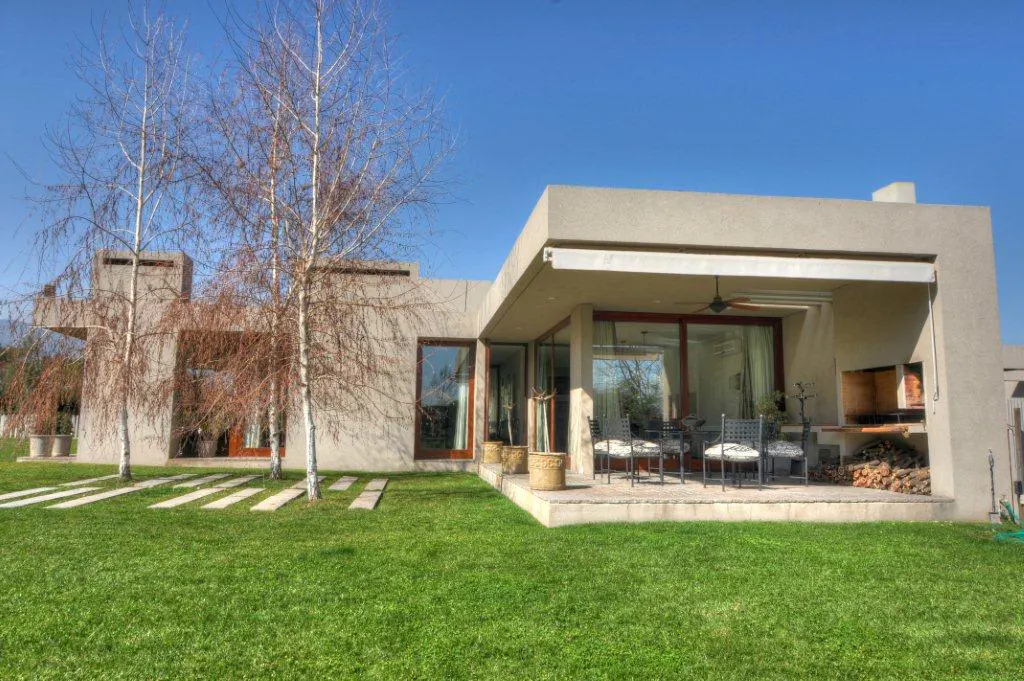 Modern beige house with a green lawn, stone path, and patio with a dining set and outdoor fireplace. Blue sky.
