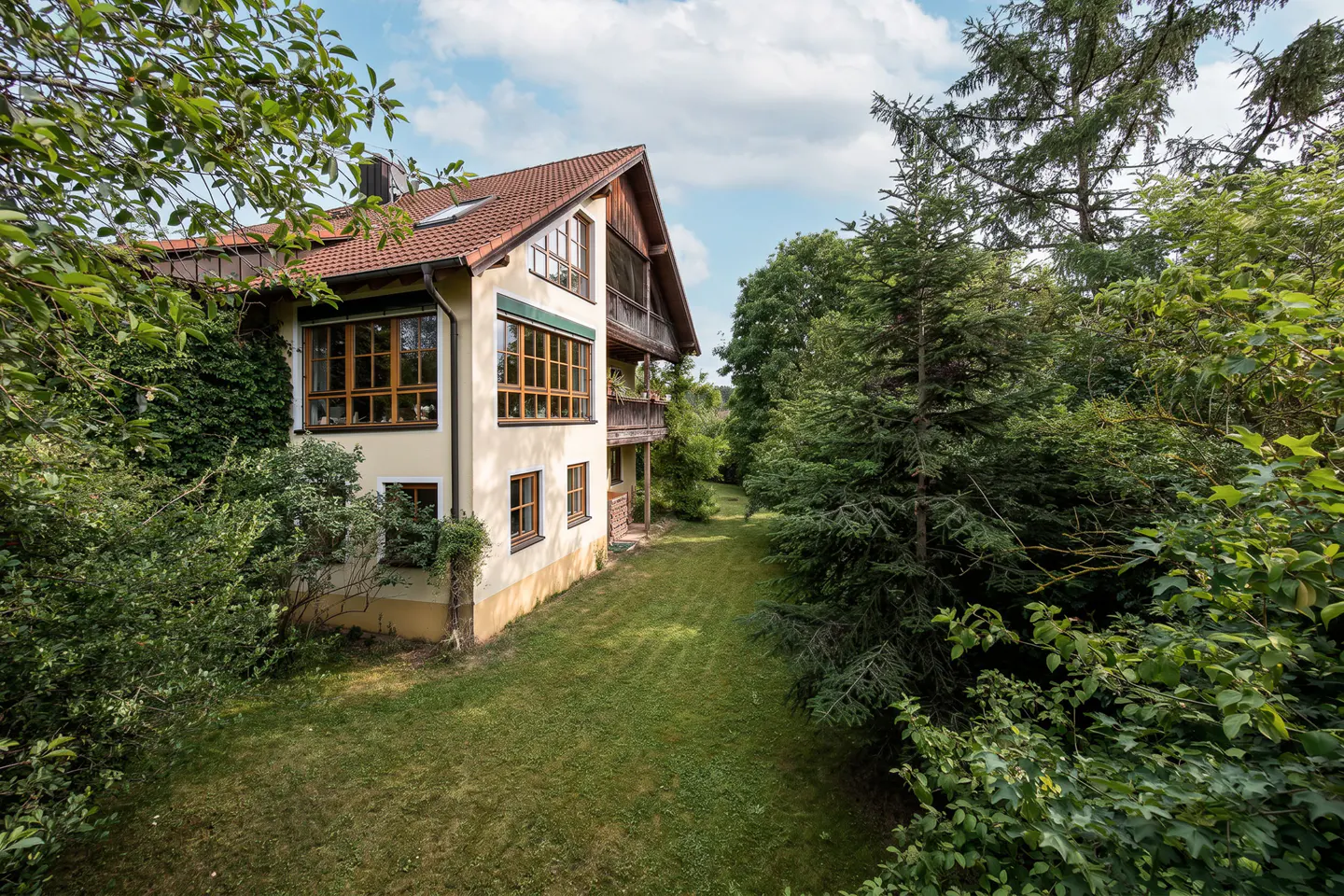 A two-story cream-colored house with a red tile roof, wooden balconies, and large windows, set in a lush, green garden.