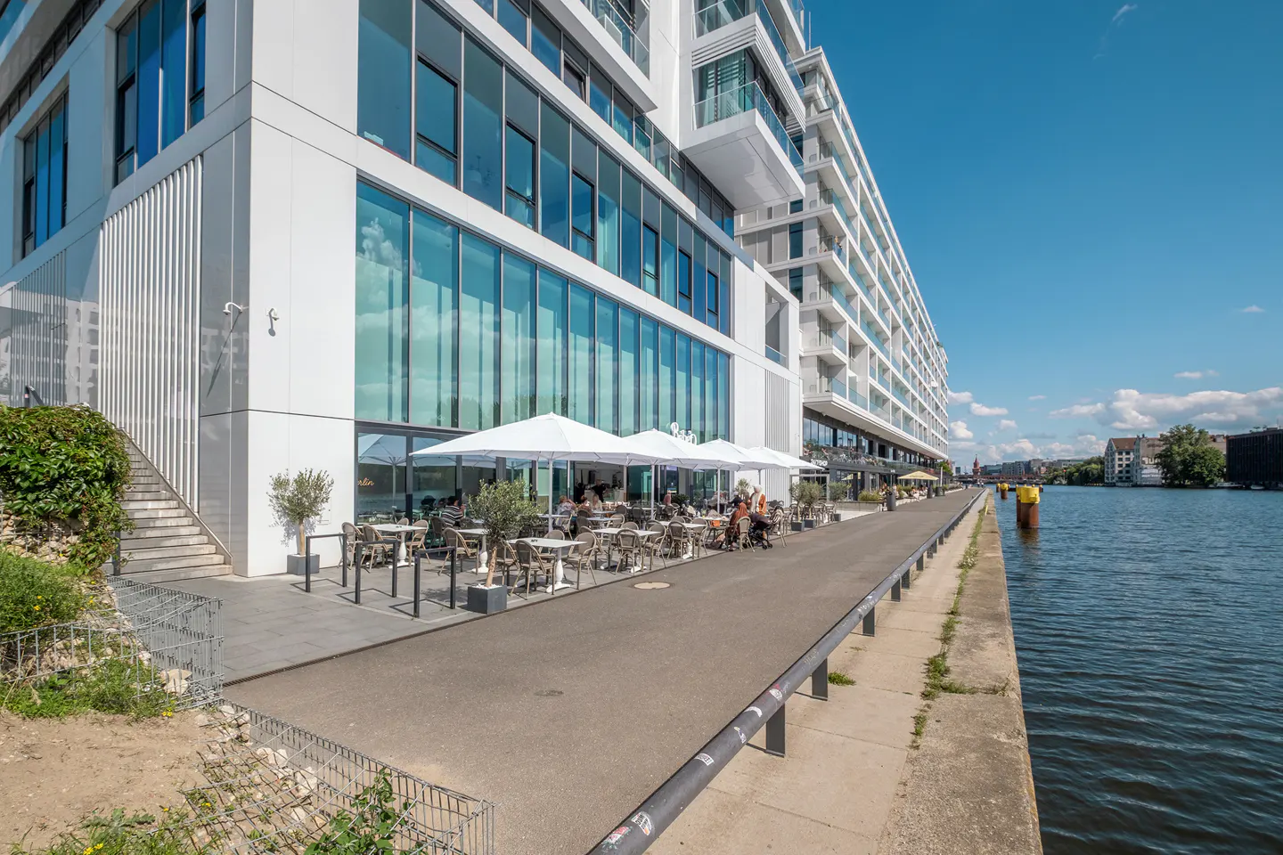 Modern white building with a waterfront cafe. People sit under white umbrellas at tables. Blue sky and water.