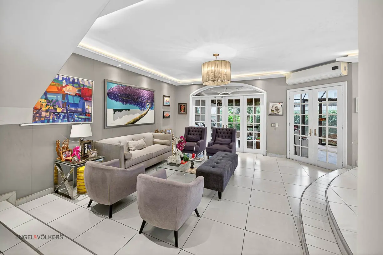 Living room with grey walls, white tile floor, sofa, chairs, ottoman, and French doors to the outside. Artwork hangs on the walls.