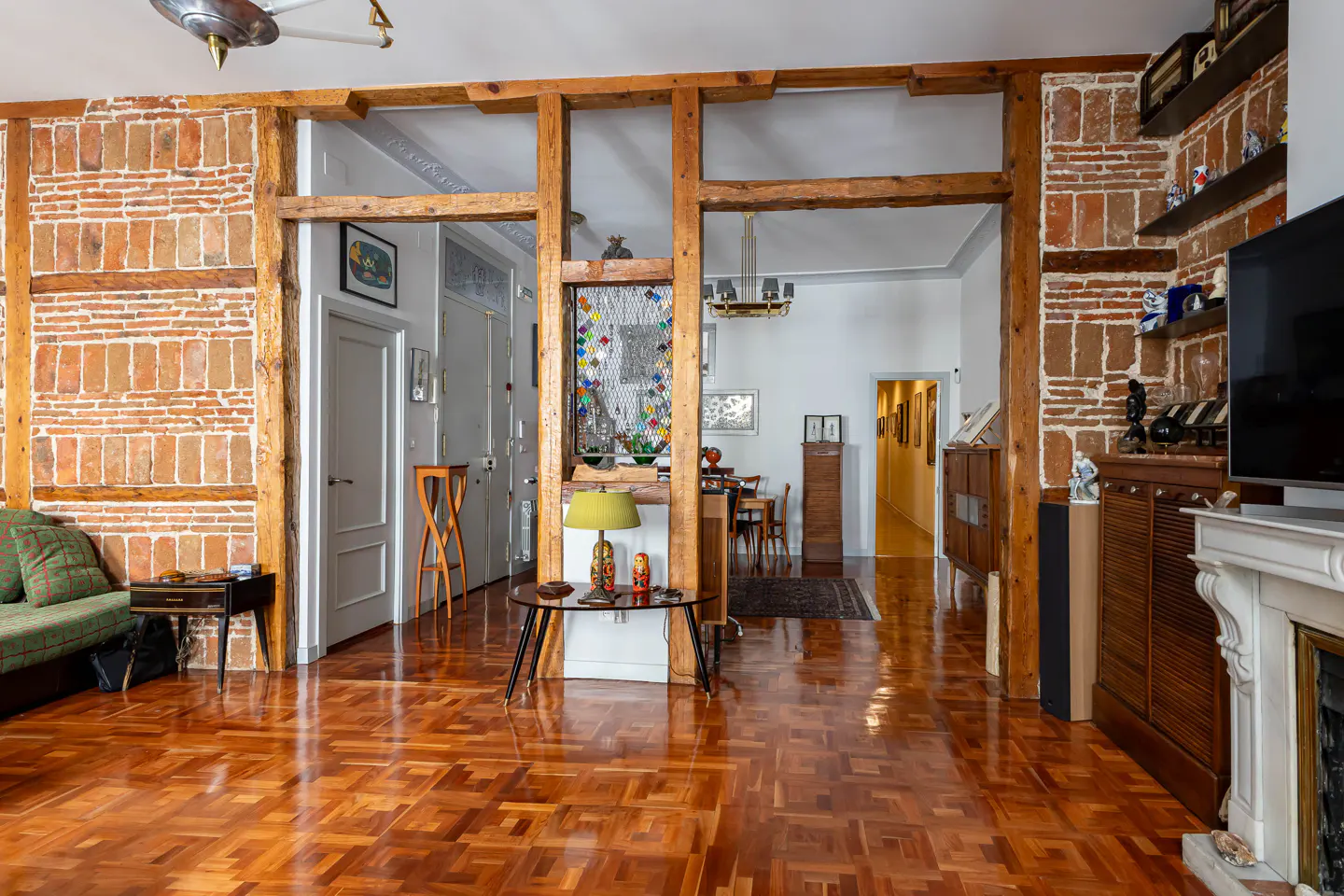 A living room with brick walls, wooden beams, and parquet floors. A green sofa is on the left, and a fireplace is on the right. A dining room is visible in the background.