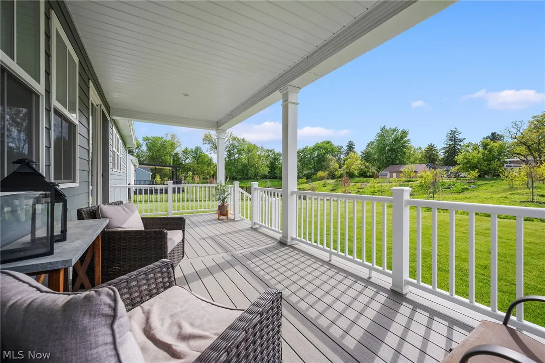 Covered porch with wicker chairs, gray cushions, and a lantern. White railing overlooks a green lawn and trees under a blue sky.