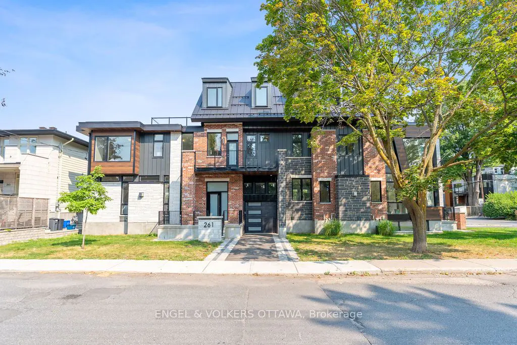 Modern townhouse with brick, stone, and black metal siding. A tree with green leaves is on the right. The address "261" is visible.