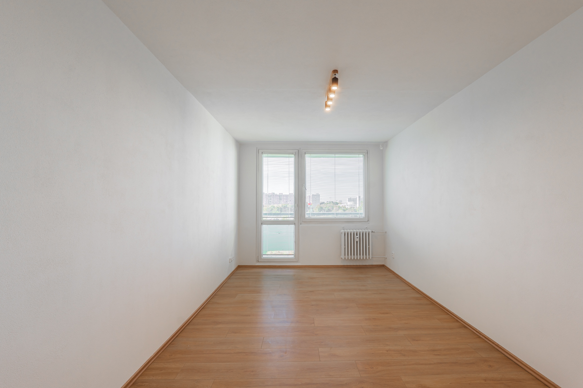 Empty room with white walls, light wood flooring, a ceiling light fixture, a window, and a white radiator. [AI-generated text]