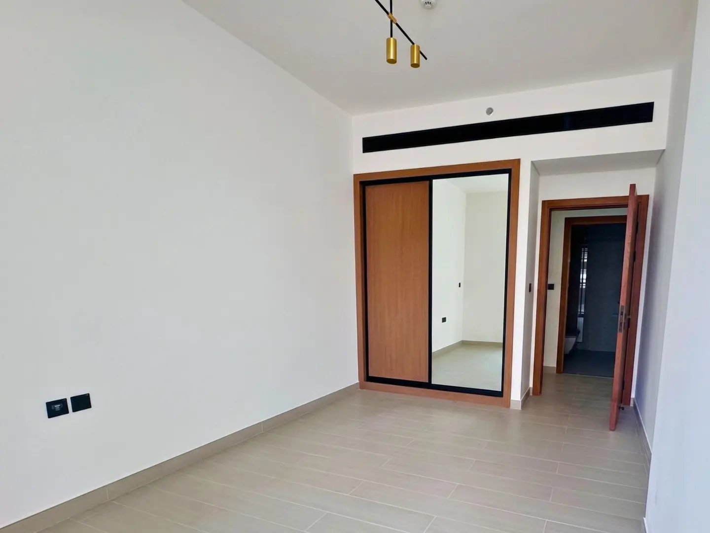 Bright, empty room with white walls, light wood-look tile floor, and a closet with sliding wood and mirrored doors. An open doorway leads to a bathroom.
