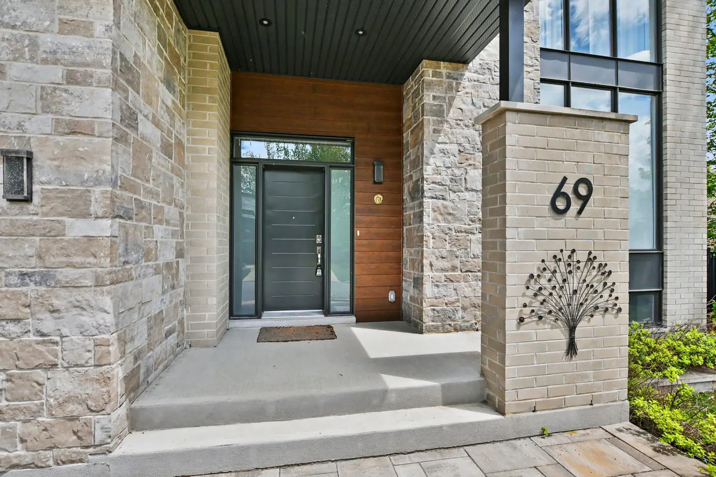 Modern home exterior with stone and wood accents. A gray front door is flanked by sidelights. Address number "69" is displayed on a brick pillar.