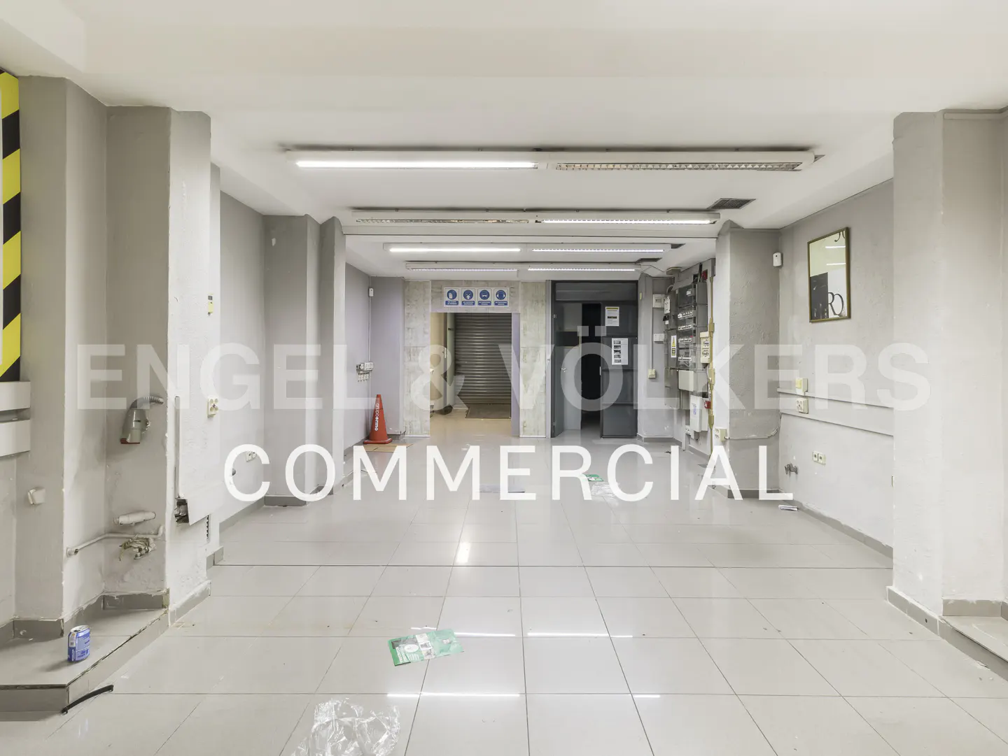 Empty commercial space with grey walls, white tiled floors, fluorescent lighting, and a rear entrance with a metal shutter.