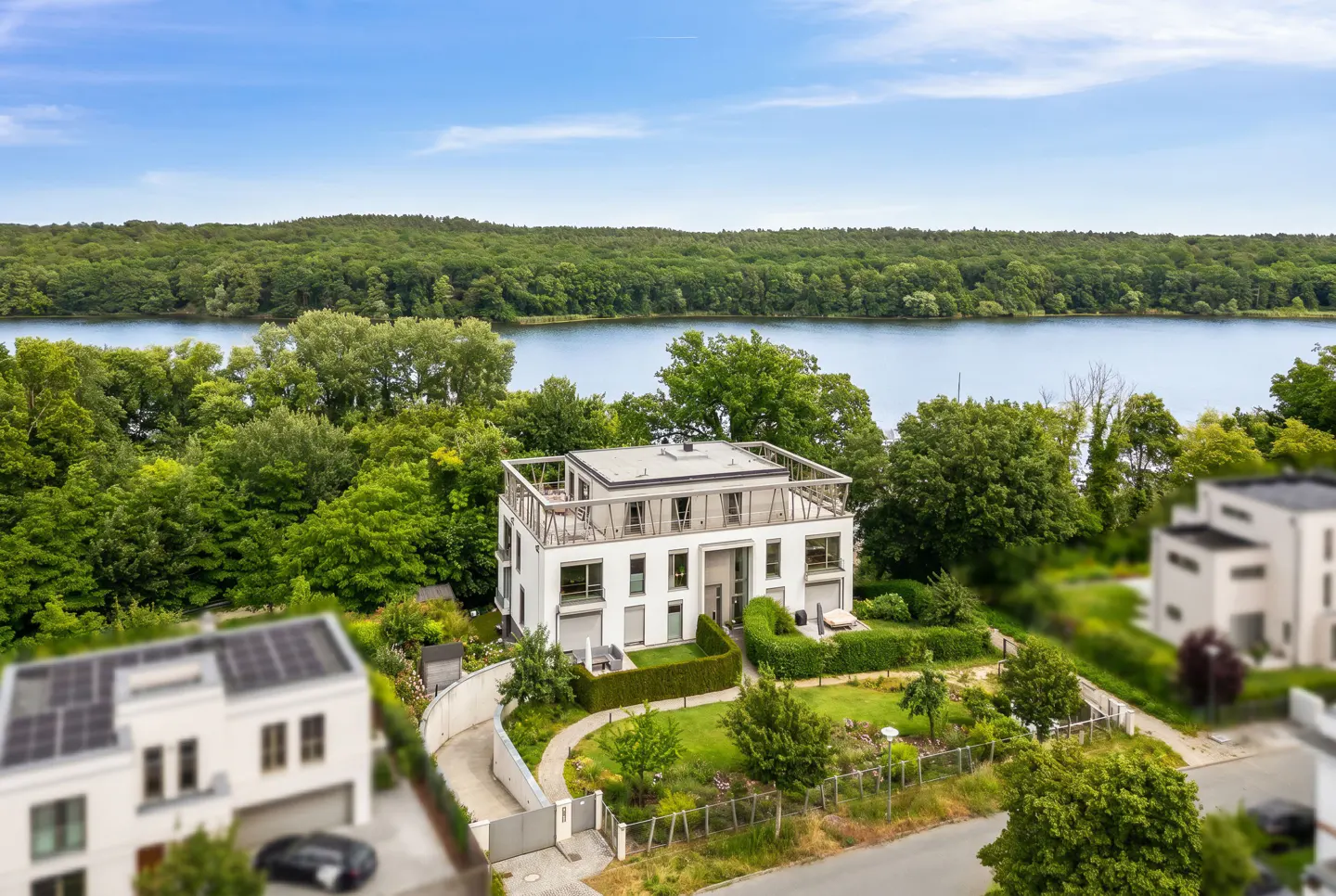 Modern white multi-story house with a rooftop terrace, surrounded by lush green trees and a lake in the background.
