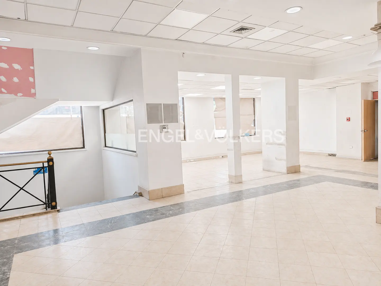 A bright, empty commercial space with white walls, tiled floors, and support columns. A staircase with a black railing is visible on the left.