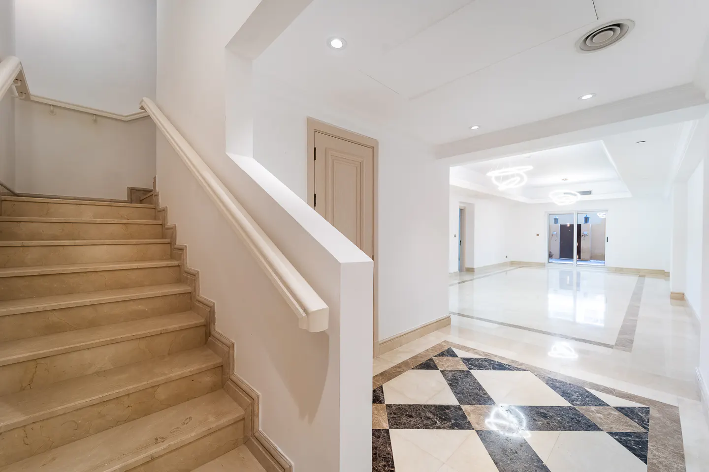 Bright foyer with marble stairs, white walls, and a black and tan checkered marble floor leading to a large, open room.