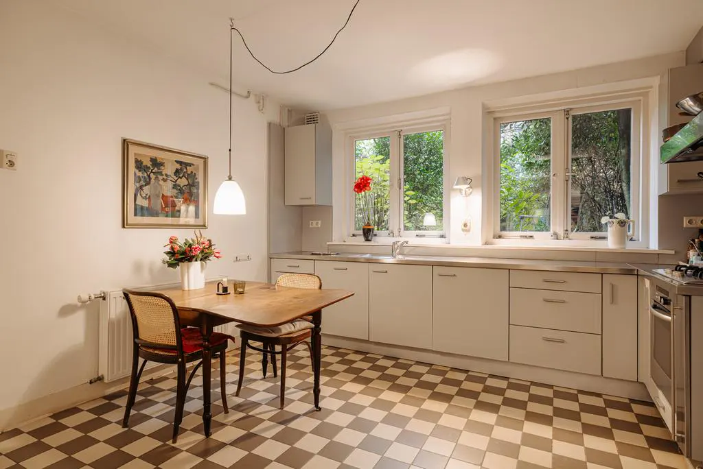 Bright kitchen with checkered floor, white cabinets, and a wooden table with flowers. Windows overlook green trees.