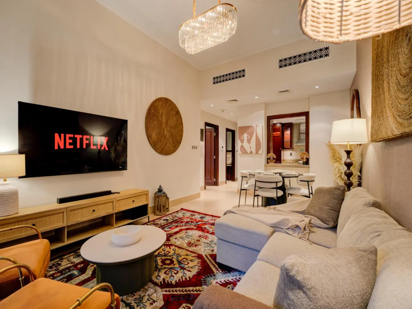 Modern living room with a large TV, beige sectional sofa, patterned red rug, and a dining area in the background.