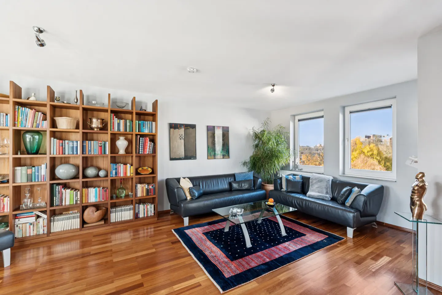 Living room with hardwood floors, a large bookcase, two black leather sofas, and a glass coffee table on a blue rug.
