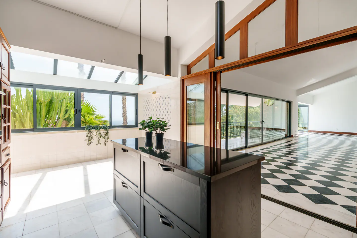 Bright, modern kitchen with black island, pendant lights, and a sunroom with large windows overlooking greenery and the ocean. Black and white checkered floors.