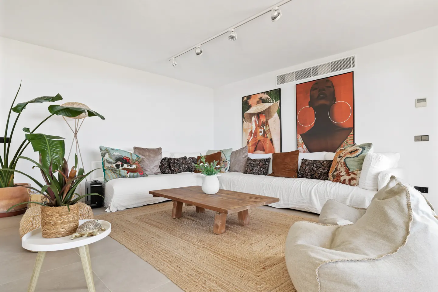 Bright living room with white walls, a large white sofa with pillows, and a wooden coffee table on a jute rug. Artwork hangs above the sofa.