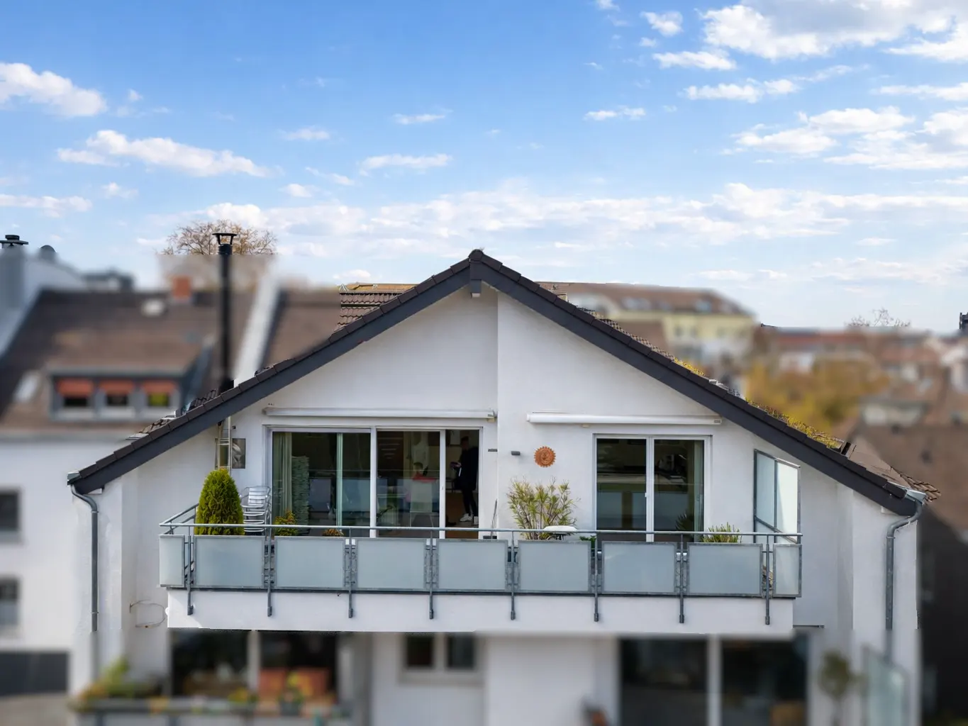 Exterior view of a white house with a balcony, sliding glass doors, and a dark gray roof against a blue sky.