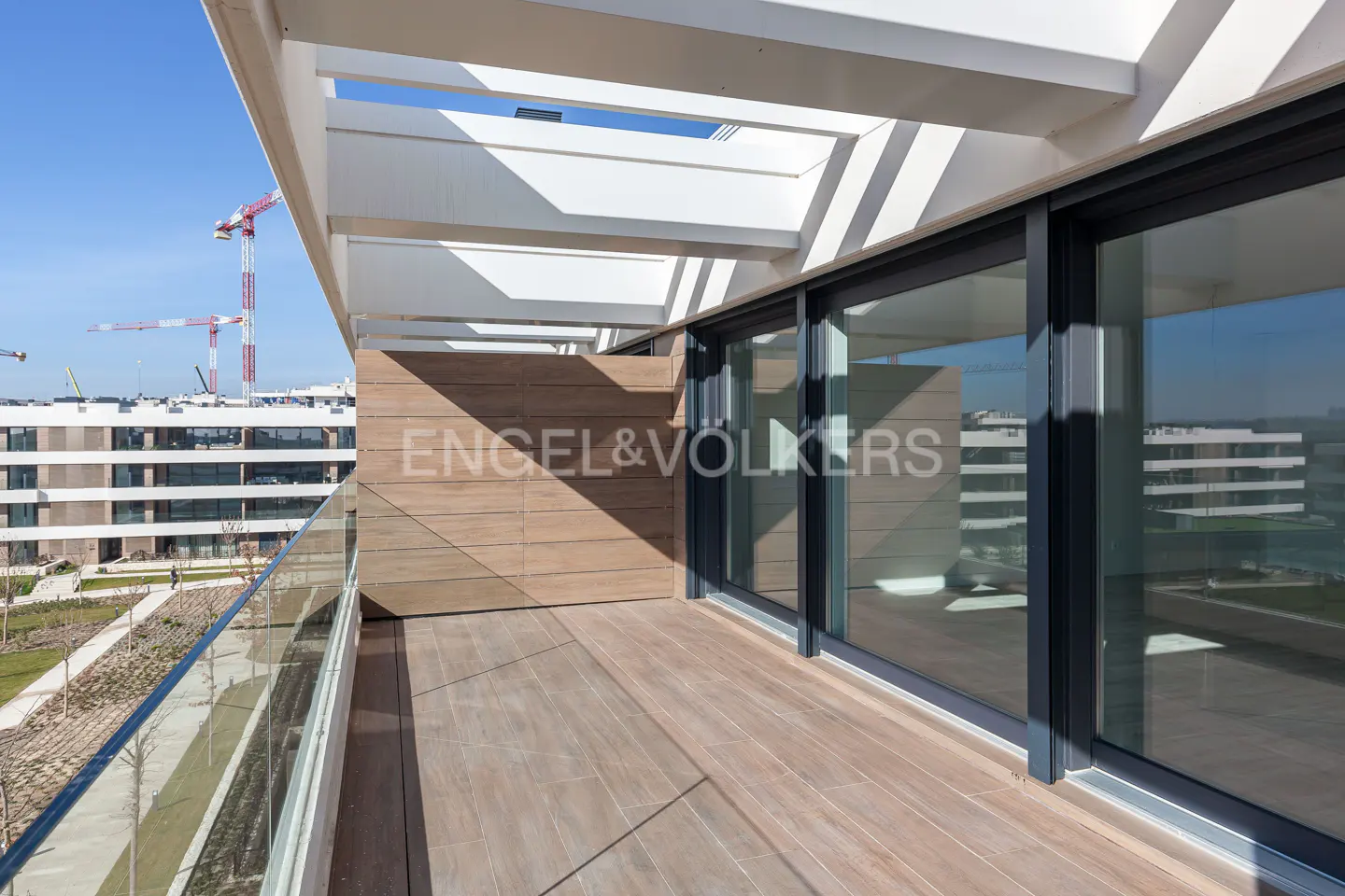 Balcony with wood floors, glass railing, and sliding glass doors. White pergola overhead. Cranes and buildings in the background.