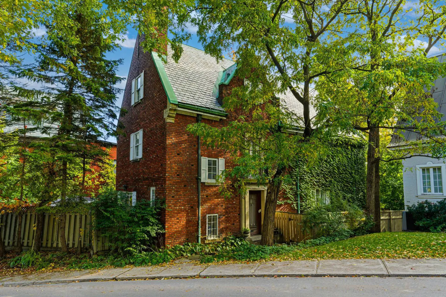 Exterior view of a two-story brick house with white shutters and a green roof, partially covered in ivy, surrounded by trees.