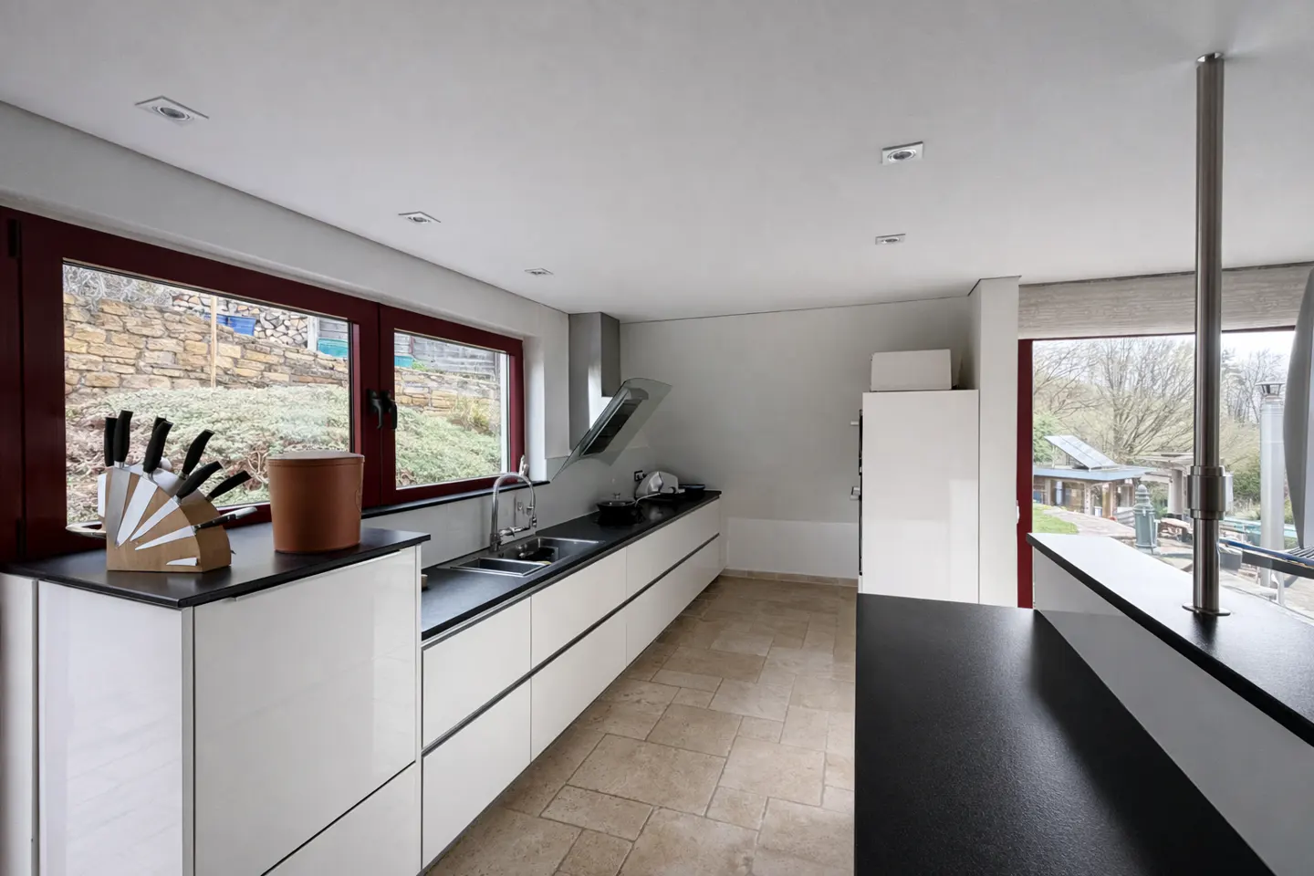 A modern kitchen with white cabinets, black countertops, and a beige tile floor. A window with a red frame looks out onto a green hillside.