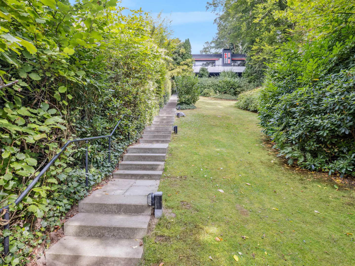Stone steps with a black handrail lead up a green lawn to a modern house surrounded by lush greenery.