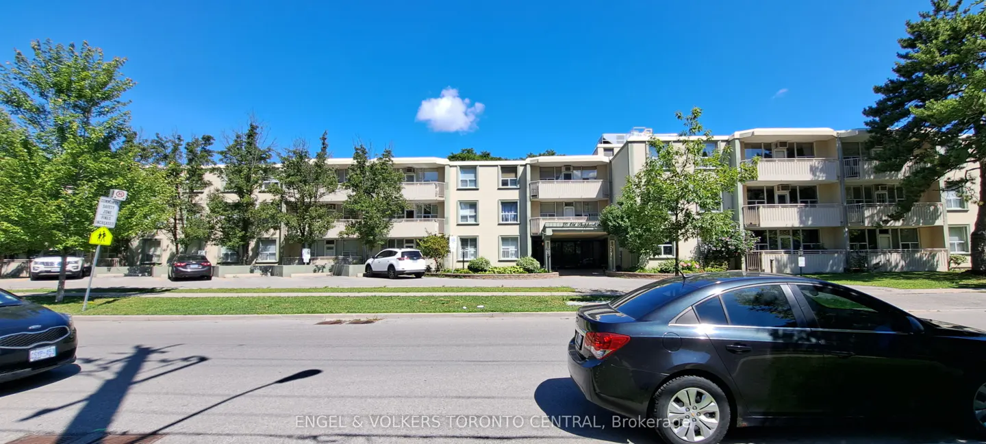 A three-story apartment building with balconies, cars parked in front, and a blue sky above.