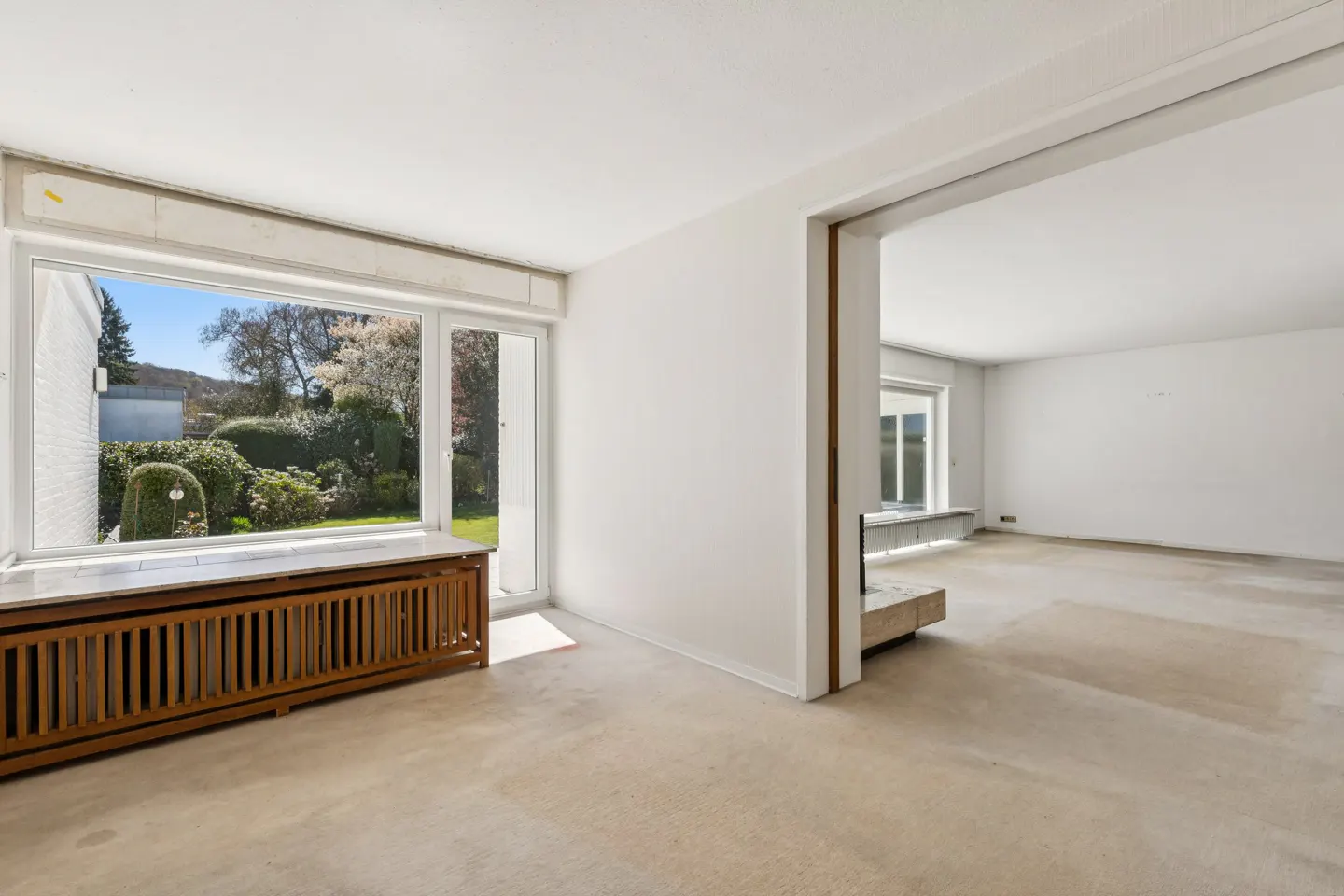 Bright, empty living room with a large window overlooking a green garden. A wooden radiator cover sits beneath the window. An open doorway leads to another room.