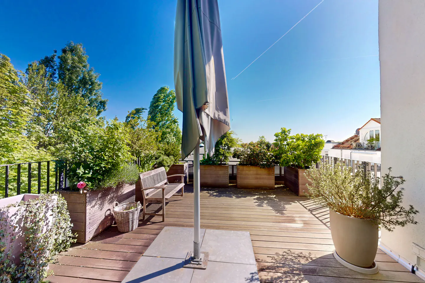 A sunny rooftop patio with wooden decking, planters with greenery, a bench, and a closed gray umbrella.