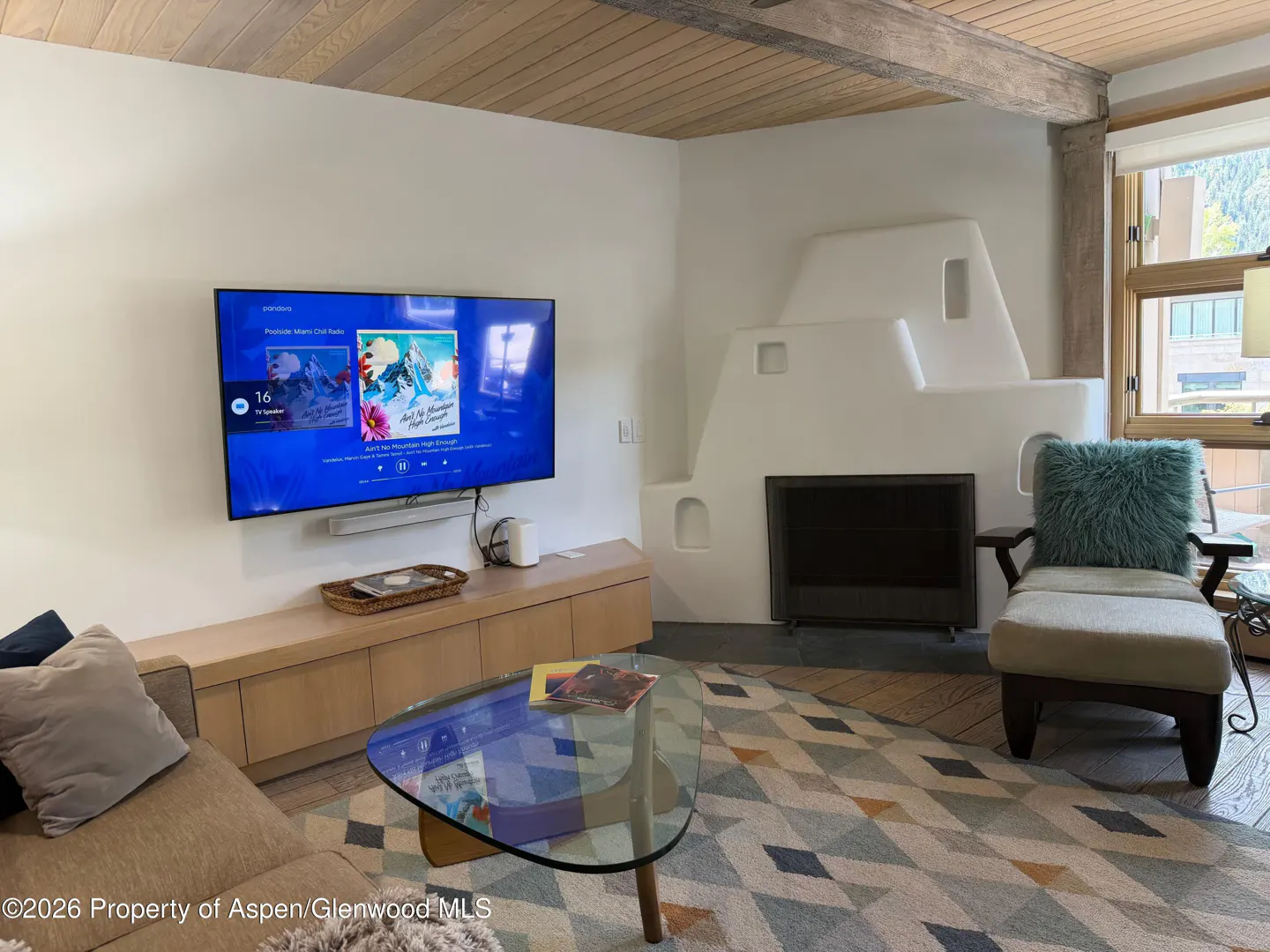 Living room with a wall-mounted TV, a white kiva-style fireplace, a glass coffee table, and a cozy armchair on a rug.