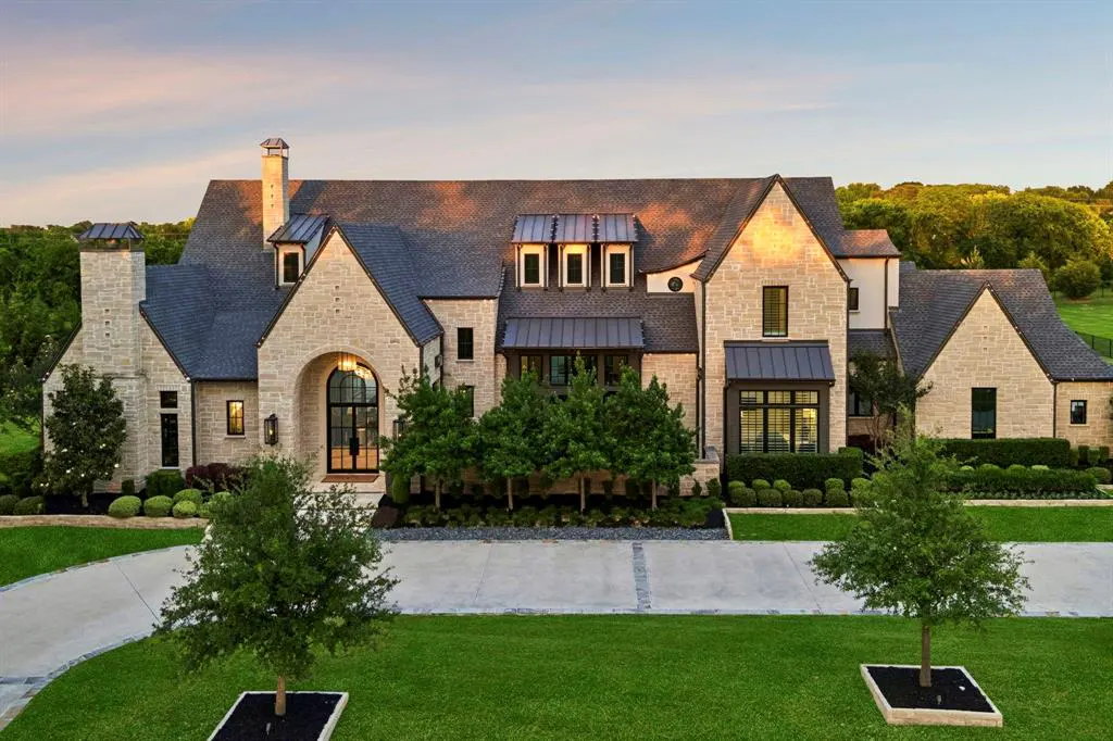 A large, two-story stone house with a dark gray roof sits on a green lawn with trees and shrubs.