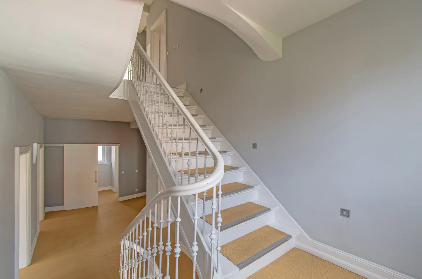 Elegant white staircase with ornate railings and light wood treads in a bright, minimalist hallway with grey walls.