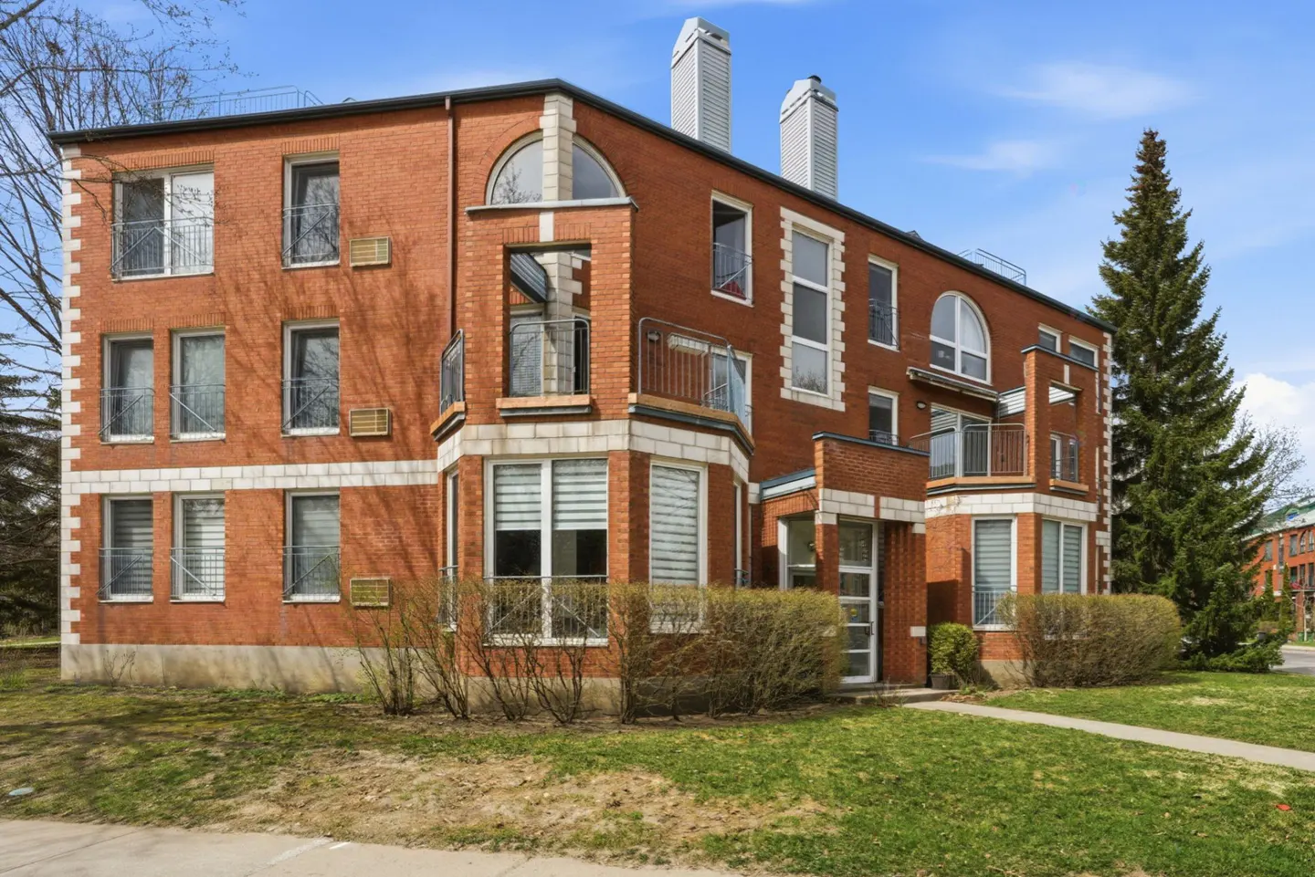 Exterior of a three-story red brick apartment building with white trim and balconies. A tall evergreen tree is on the right.
