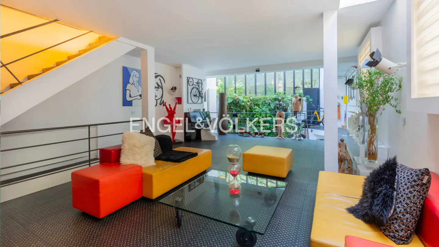 Bright, modern living room with red and yellow modular seating, glass table, and yellow stairs. Artwork adorns the white walls. Engel & Völkers logo visible.