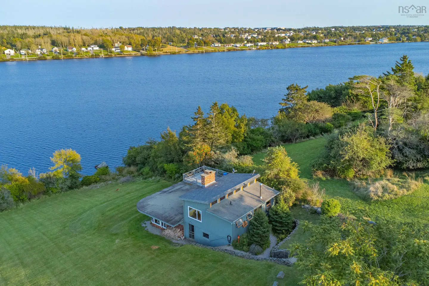 Aerial view of a blue house with a chimney, surrounded by green grass and trees, overlooking a wide blue river.