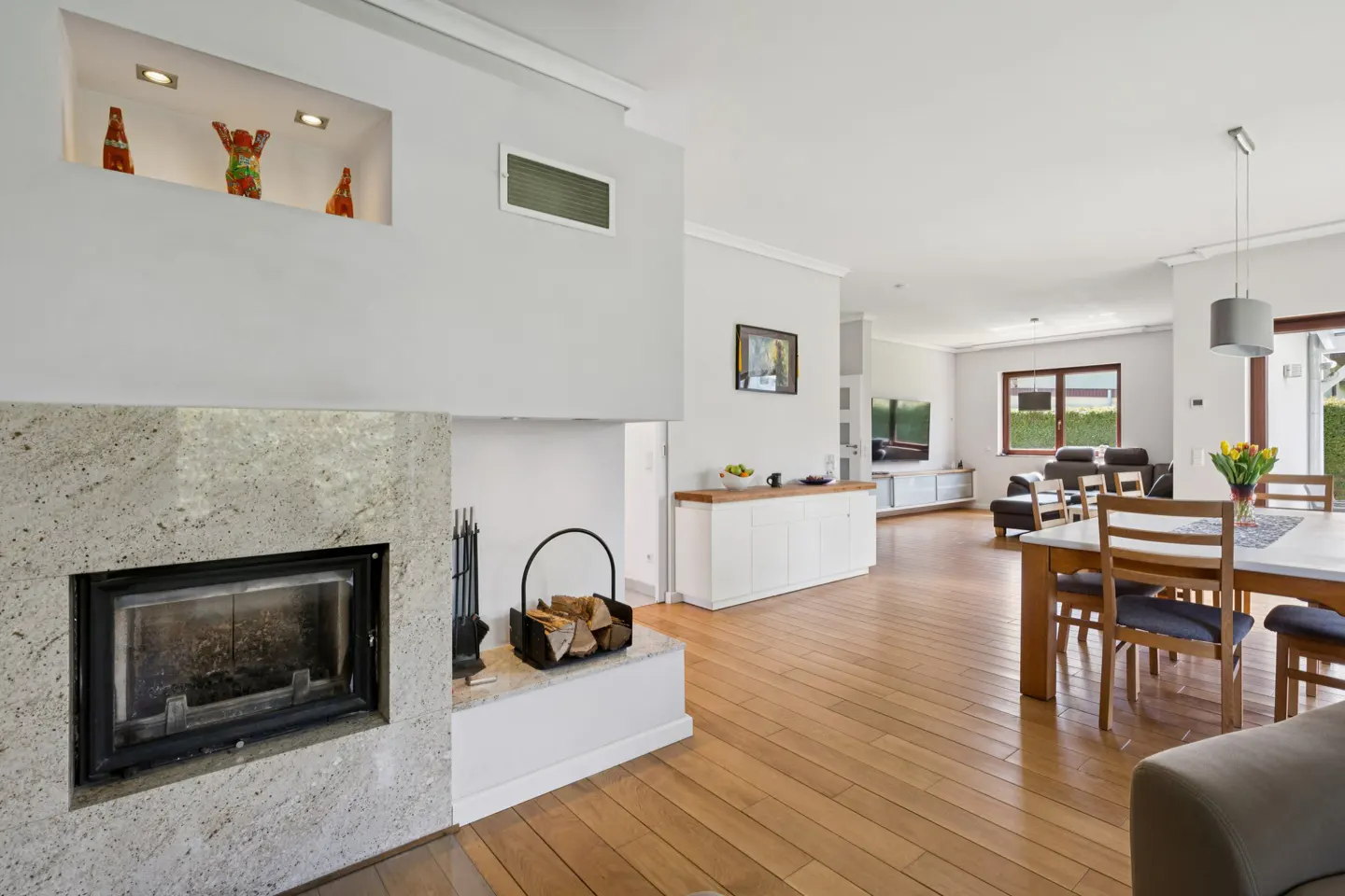 Bright, open-concept living space with hardwood floors. A stone fireplace anchors the room, with a dining area and living room visible in the background.