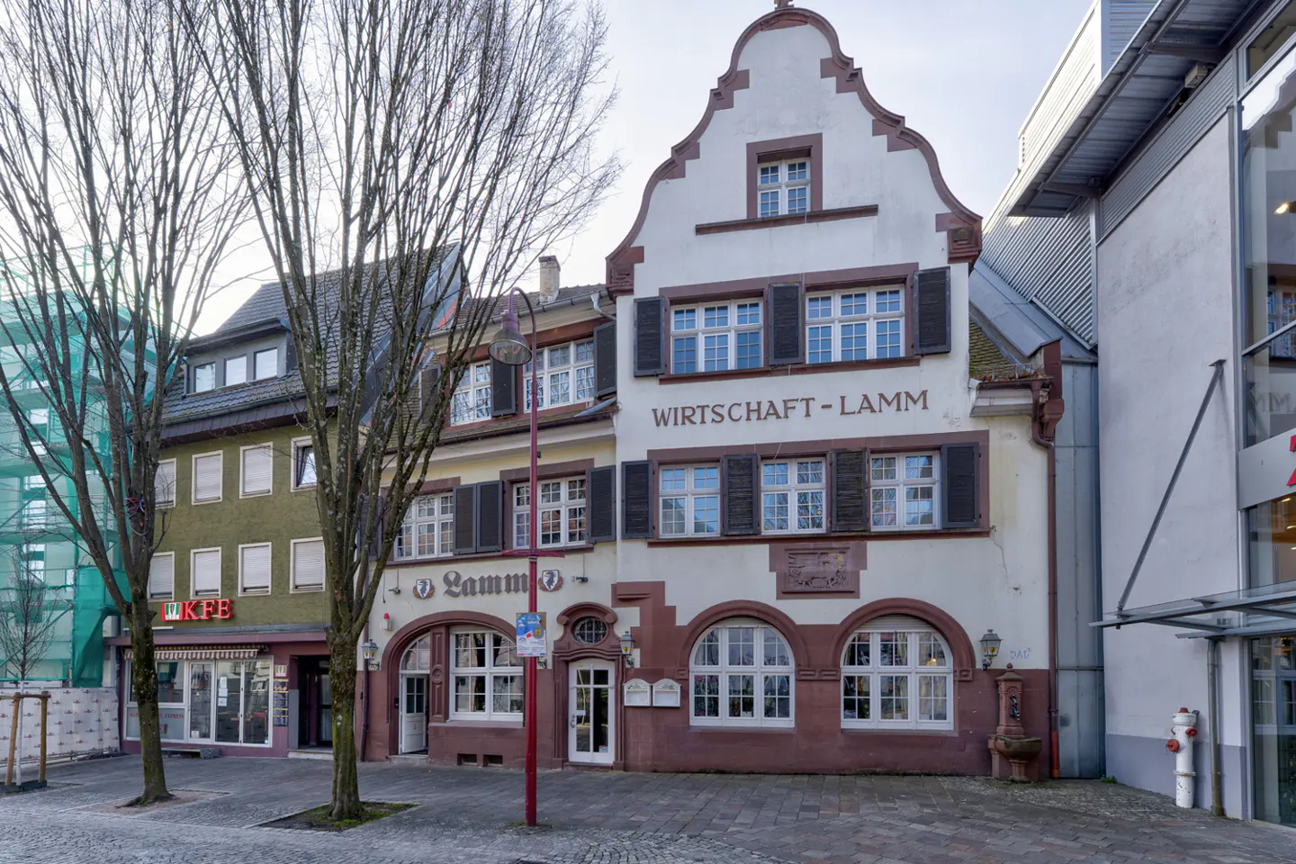 Exterior view of "Wirtschaft-Lamm" building with brown shutters and arched windows, next to a green building and modern structure.