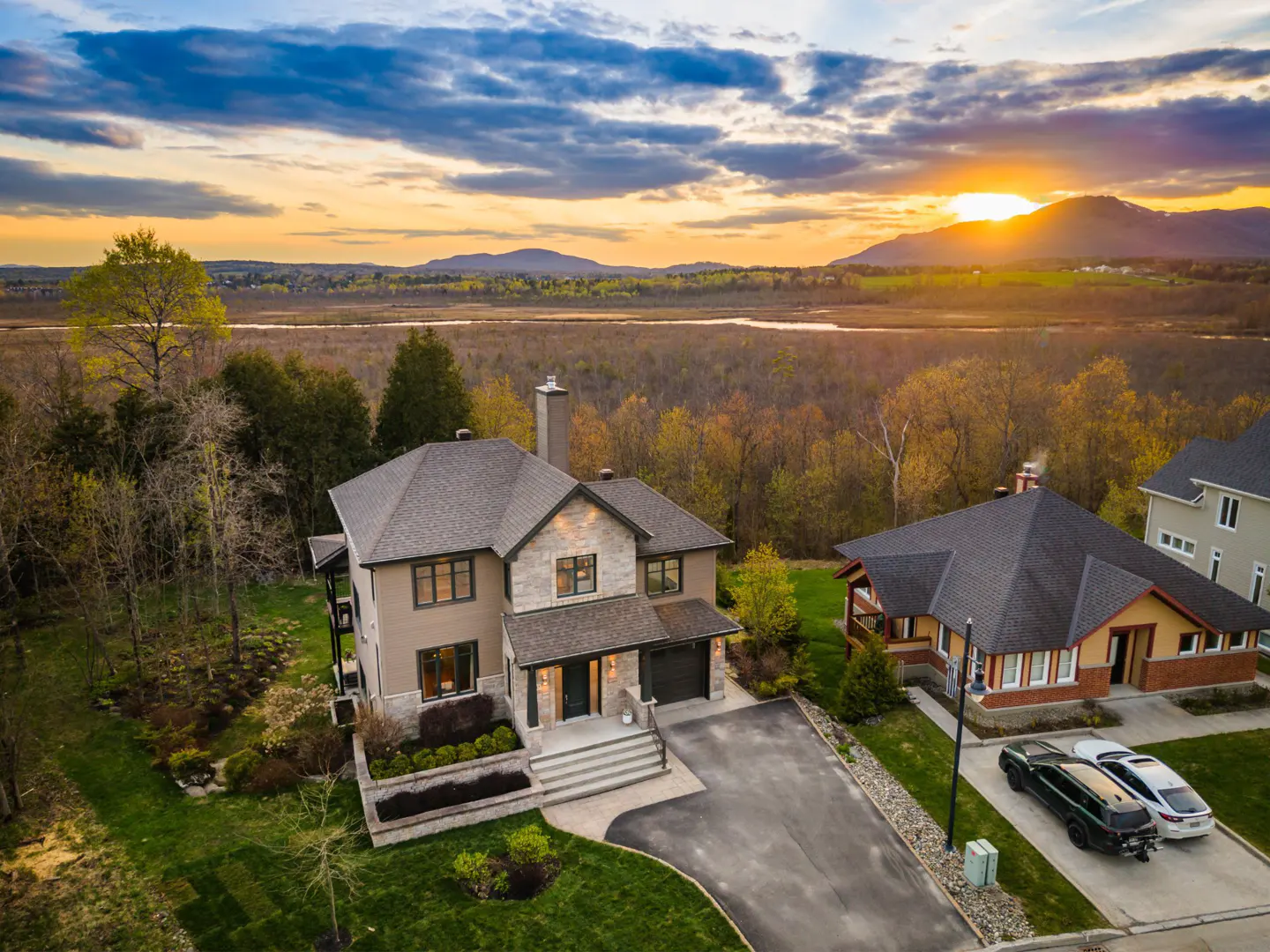 Aerial view of a two-story beige house with a gray roof, a driveway, and a green lawn at sunset. Mountains in the background.