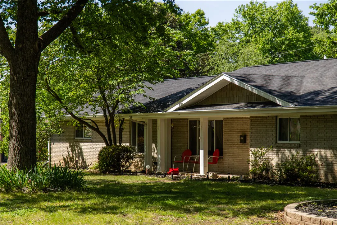 A ranch-style brick house with a dark roof and a covered porch with red chairs. Green trees surround the house.