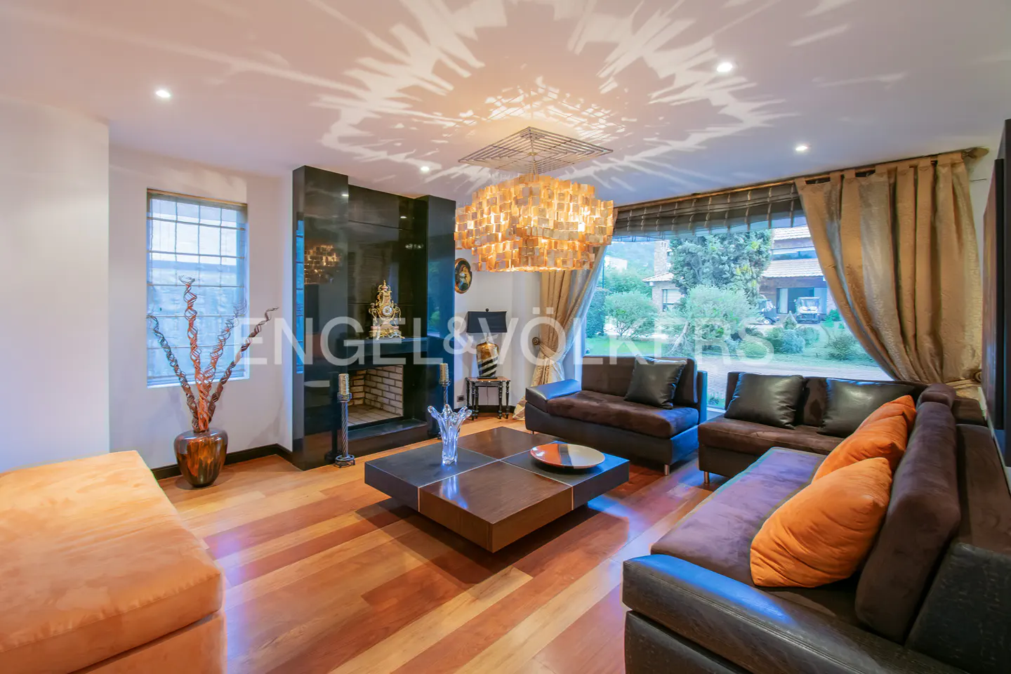 Living room with hardwood floors, brown sofas, and a black marble fireplace. A large window overlooks a green lawn.