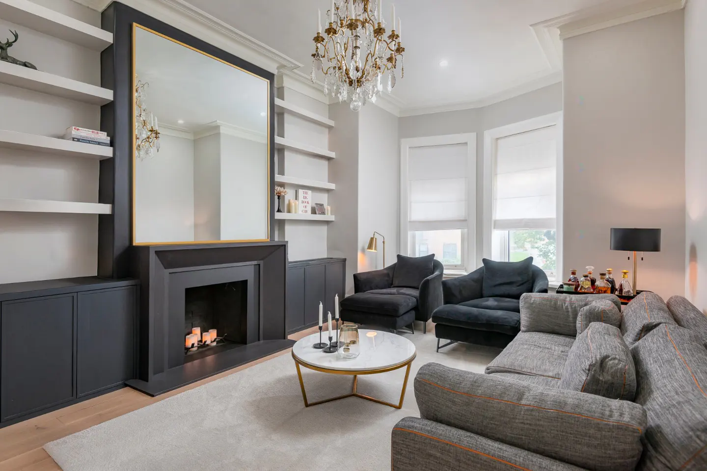 Living room with gray sofa, black chairs, and fireplace with a large mirror. A chandelier hangs from the ceiling.
