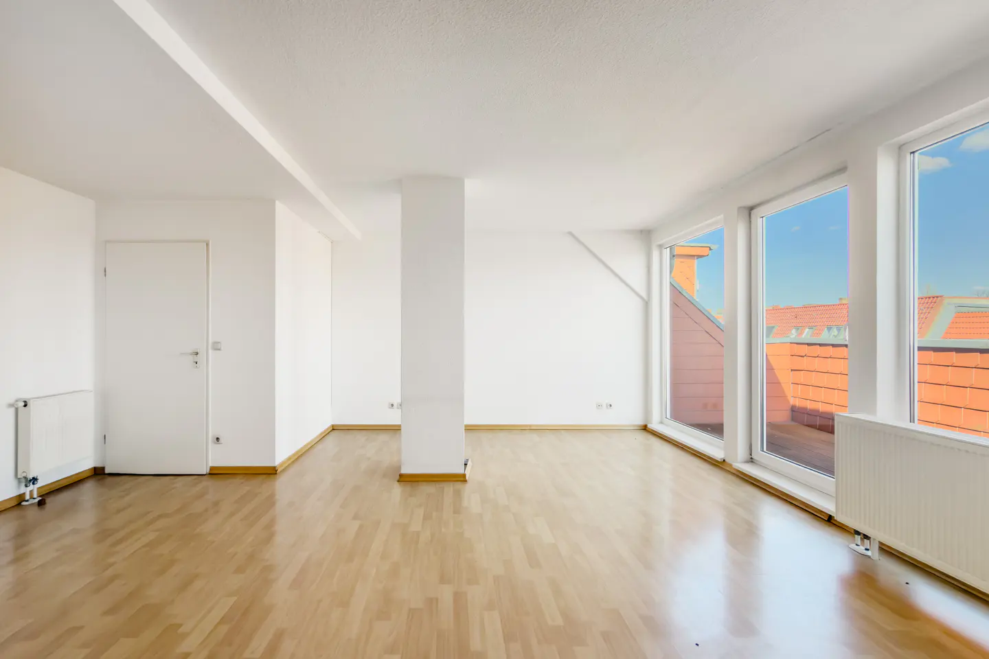 Bright, empty room with wood floors, white walls, and large windows showing rooftops and blue sky.