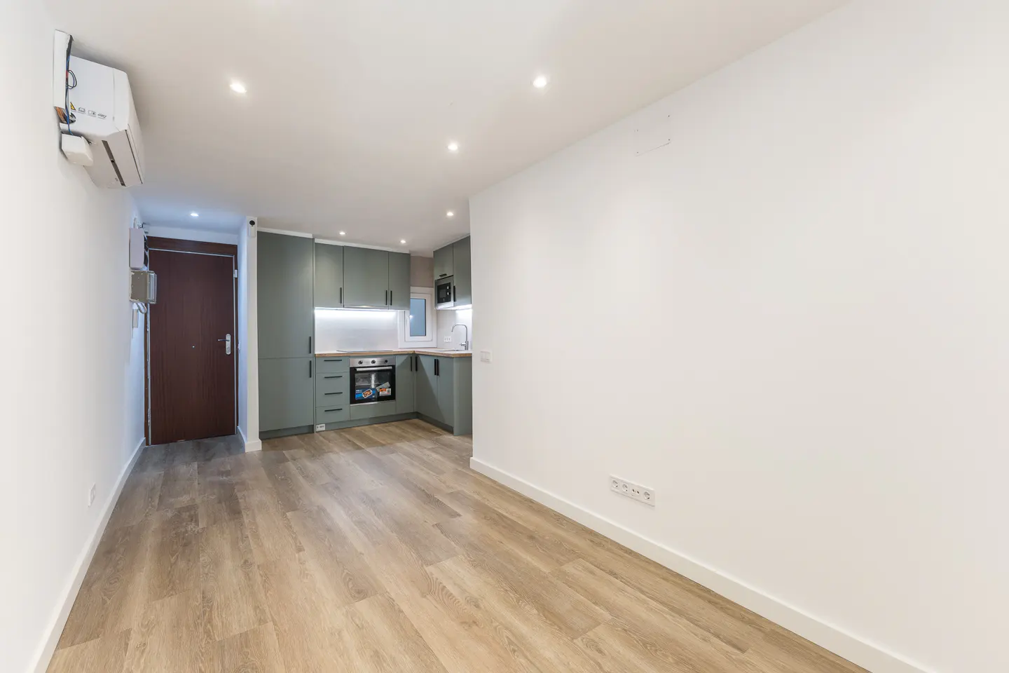 A bright, empty apartment with wood floors, white walls, and a green kitchen.