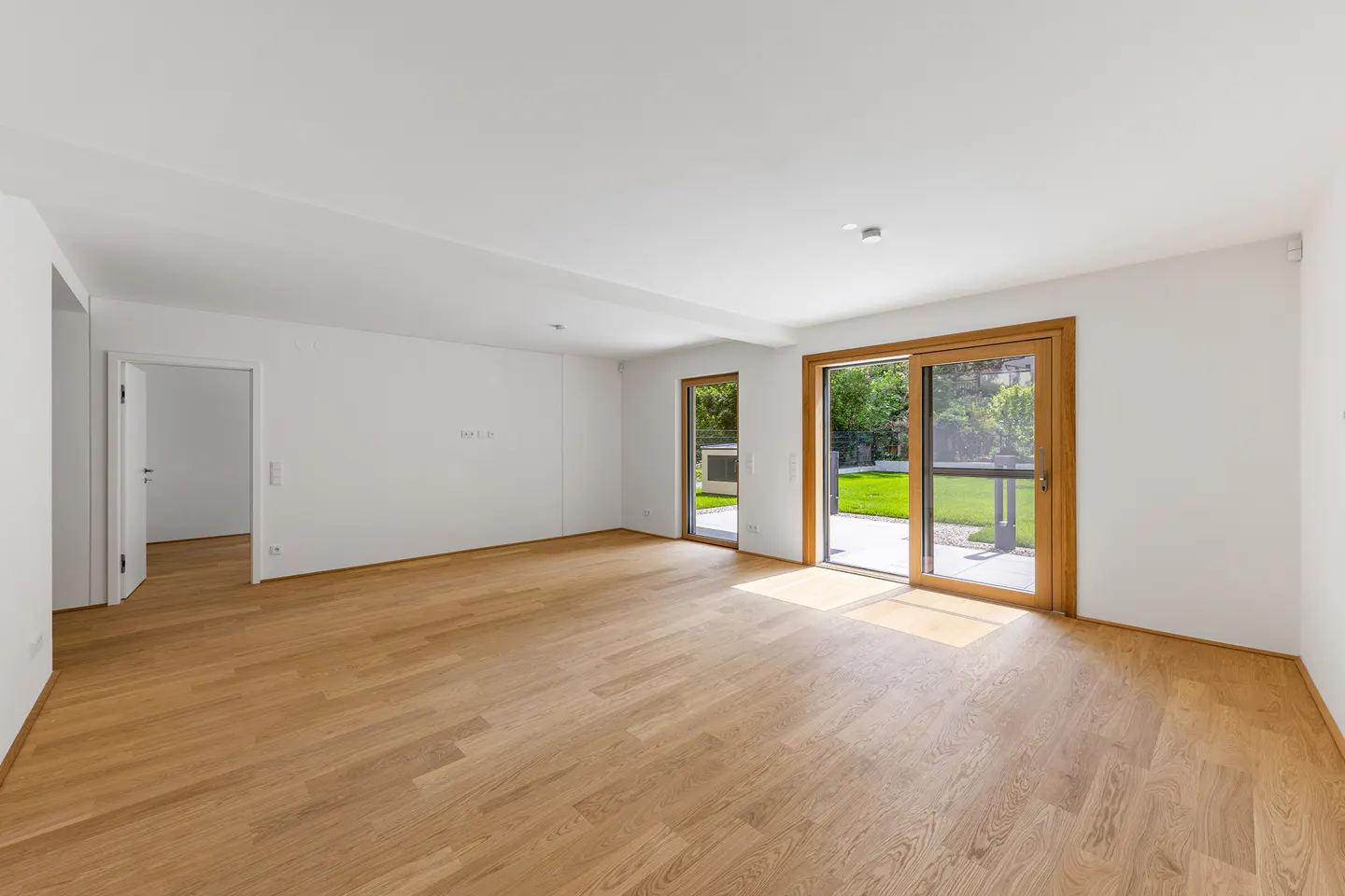 Bright, empty room with wood floors, white walls, and sliding glass doors to a green lawn. A doorway leads to another room.