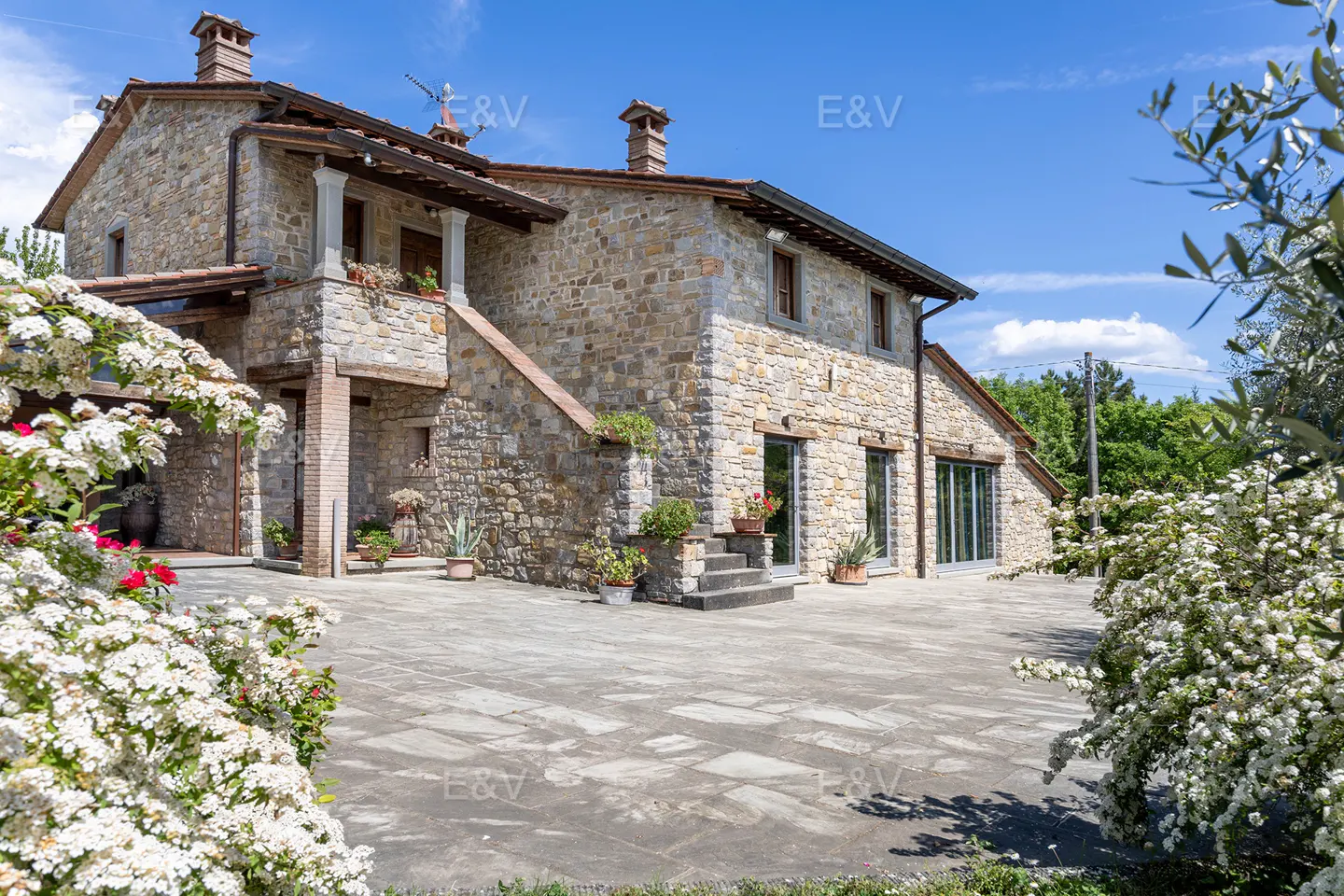 Exterior view of a two-story stone house with a tiled roof, a stone patio, and white flowering bushes.